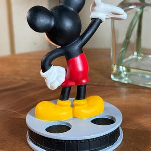 May include: A Mickey Mouse figurine in a classic pose, with black ears and head, red shorts, yellow shoes, and white gloves. The figure stands on a grey film reel base. A decorative item.