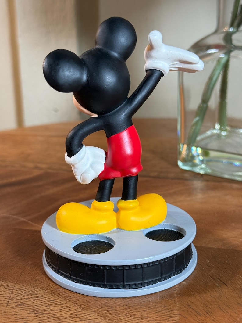 May include: A Mickey Mouse figurine in a classic pose, with black ears and head, red shorts, yellow shoes, and white gloves. The figure stands on a grey film reel base. A decorative item.