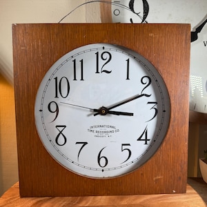 Puede incluir: Un reloj cuadrado con marco de madera y esfera blanca con números negros. La esfera del reloj tiene el texto "INTERNATIONAL TIME RECORDING CO.". Las manecillas del reloj señalan aproximadamente las 3:10. Un reloj vintage está en el fondo.