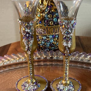 K&ouml;nnte beinhalten: Zwei goldbemalte Schnapsgl&auml;ser mit einem dekorativen Design aus schimmernden lila und wei&szlig;en Strasssteinen. Die Gl&auml;ser stehen auf einem goldenen Tablett.
