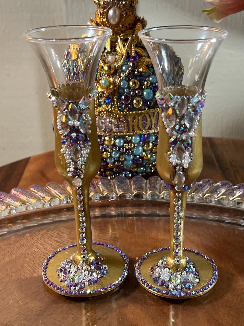 K&ouml;nnte beinhalten: Zwei goldbemalte Schnapsgl&auml;ser mit einem dekorativen Design aus schimmernden lila und wei&szlig;en Strasssteinen. Die Gl&auml;ser stehen auf einem goldenen Tablett.