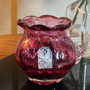 May include: A cranberry-colored glass pitcher with a ruffled rim and a clear handle. The pitcher has a round body and a label that reads "Pilgrim Cranberry Glass". The pitcher is likely vintage and is a decorative item.