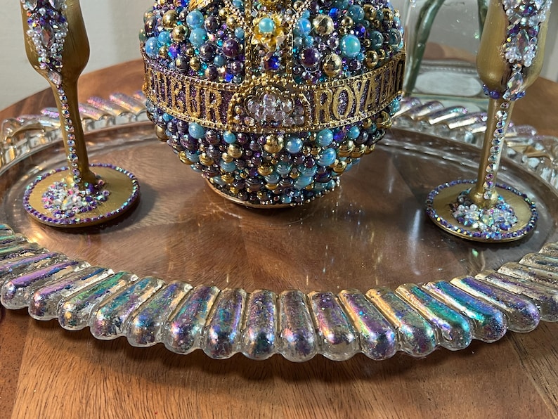 K&ouml;nnte beinhalten: Ein Serviertablett aus Glas mit irisierendem Finish und einem perlenbesetzten, goldverzierten Ornament in der Mitte. Zwei goldbemalte Gl&auml;ser mit Strasssteinen befinden sich auf dem Tablett.