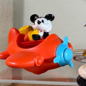 May include: A red plastic toy airplane with a blue propeller. A Mickey Mouse figure is seated in the cockpit, wearing a yellow and red outfit. The toy is a collectible item.