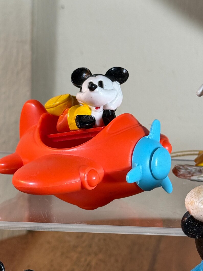 May include: A red plastic toy airplane with a blue propeller. A Mickey Mouse figure is seated in the cockpit, wearing a yellow and red outfit. The toy is a collectible item.