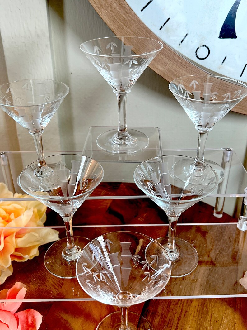 Op de afbeelding: Set van zes heldere glazen martini glazen met gegraveerde bamboe motieven. De glazen zijn gerangschikt op een heldere acryl trapstandaard.