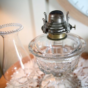 May include: A clear glass oil lamp with a metal burner and a decorative glass shade. The lamp is sitting on a white lace tablecloth.