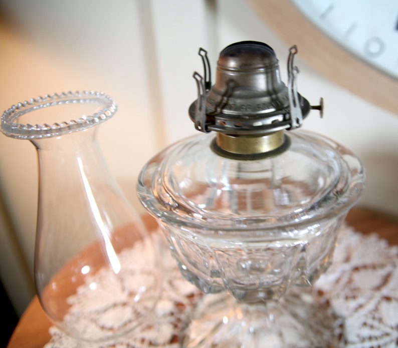 May include: A clear glass oil lamp with a metal burner and a decorative glass shade. The lamp is sitting on a white lace tablecloth.