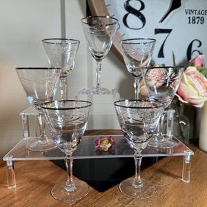 May include: A collection of clear crystal wine glasses with a silver rim and etched floral design. The glasses are arranged on clear acrylic risers, with a vintage clock and flowers in the background. The glasses are for serving wine.