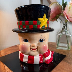 May include: A ceramic snowman-shaped container with a black top hat, a red and green checkered band, and a yellow star. The snowman has rosy cheeks, blue eyes, and a red and white striped scarf. The container is on a wooden surface.