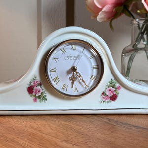 May include: A white ceramic mantel clock with a gold-rimmed circular face featuring Roman numerals and the text Formalities by Baum Bros. The clock body is arched and decorated with two floral clusters of pink and red roses on each side.