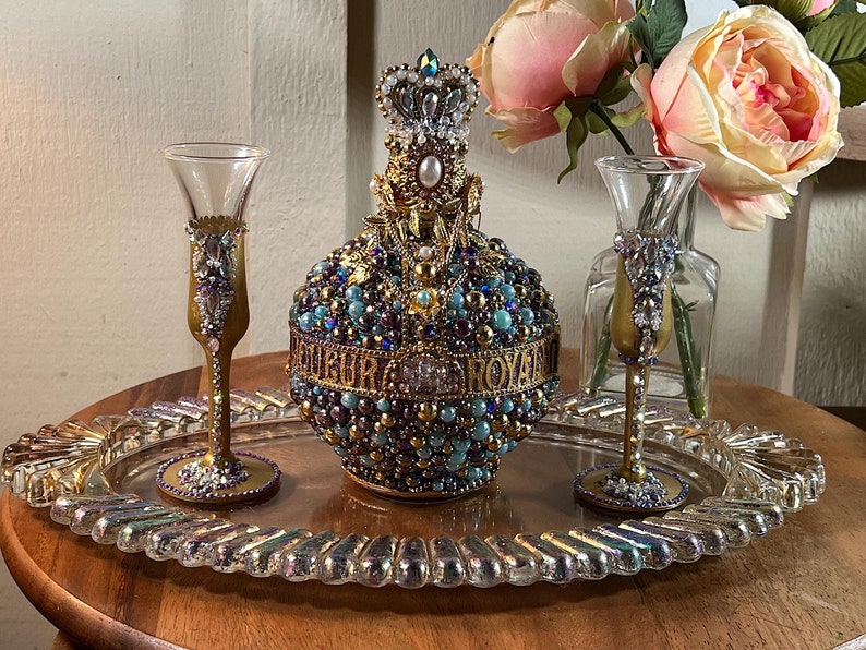 K&ouml;nnte beinhalten: Eine goldene und blaue Dekorationsflasche mit einer Krone oben, umgeben von zwei goldenen und blauen Schnapsgl&auml;sern auf einem durchsichtigen Tablett. Die Flasche und die Gl&auml;ser sind mit Perlen und Juwelen verziert.