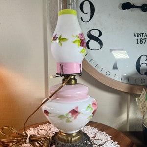 May include: A white glass lamp with pink and green floral designs. The lamp has a round base and a decorative metal stand with three legs.