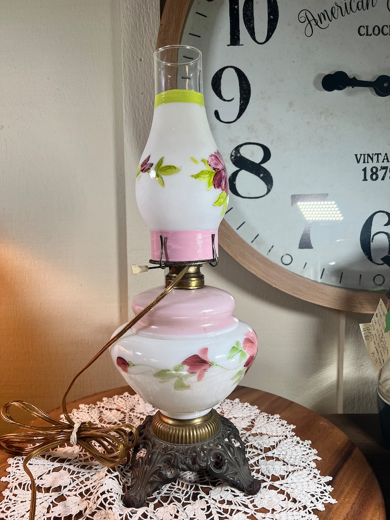 May include: A white glass lamp with pink and green floral designs. The lamp has a round base and a decorative metal stand with three legs.