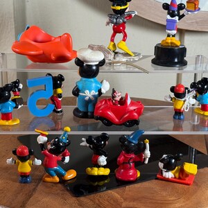 May include: A collection of Mickey Mouse figurines displayed on a clear, tiered shelf. The figurines depict Mickey in various outfits and poses, including a chef, a magician, and driving a red car. Other items include a red airplane and the number 5.