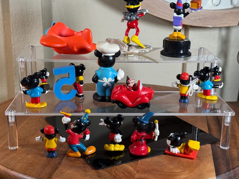 May include: A collection of Mickey Mouse figurines displayed on a clear, tiered shelf. The figurines depict Mickey in various outfits and poses, including a chef, a magician, and driving a red car. Other items include a red airplane and the number 5.