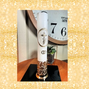 Puede incluir: Vela de boda blanca con una cruz dorada y dos anillos dorados. La vela está sobre un soporte de metal dorado.