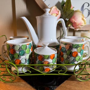 Può includere: Un servizio da caffè in ceramica bianca con una teiera, una zuccheriera e quattro tazze, tutte decorate con un motivo a bolle colorate. Il set è appoggiato su un carrello di servizio in metallo verde. Lo sfondo comprende fiori e candele.
