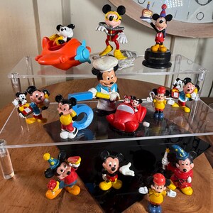 May include: A collection of Mickey Mouse figurines in various poses and outfits. The figurines are brightly colored, featuring Mickey in different costumes, including a chef, pilot, and birthday celebration attire. Some figurines are on a clear display shelf.