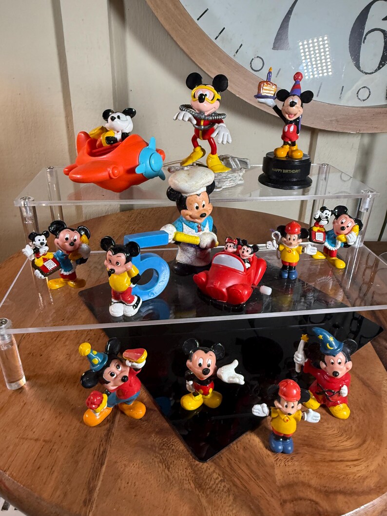 May include: A collection of Mickey Mouse figurines in various poses and outfits. The figurines are brightly colored, featuring Mickey in different costumes, including a chef, pilot, and birthday celebration attire. Some figurines are on a clear display shelf.
