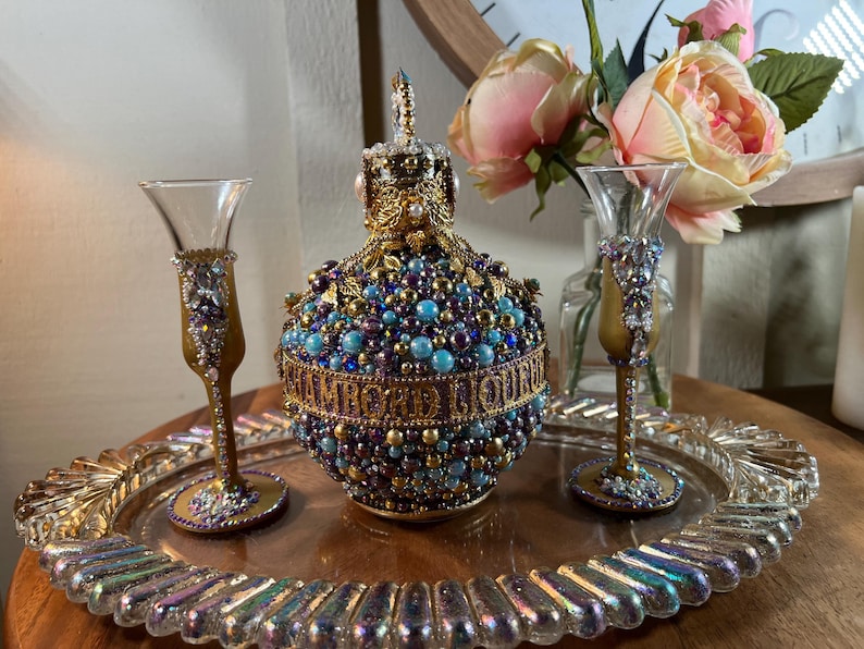K&ouml;nnte beinhalten: Ein dekoratives Tablett mit zwei goldger&auml;nderten Gl&auml;sern und einer mit Juwelen besetzten Flasche. Die Flasche ist mit blauen, violetten und goldenen Perlen verziert und hat eine goldene Krone oben. Das Tablett hat eine schillernde Oberfl&auml;che.