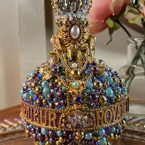 K&ouml;nnte beinhalten: Eine goldene und violette, mit Juwelen besetzte Flasche mit einem kronenf&ouml;rmigen Verschluss. Die Flasche ist mit Perlen, Strasssteinen und goldenen Akzenten verziert. Der Text "OUR ROYAL" ist um die Flasche herum geschrieben.