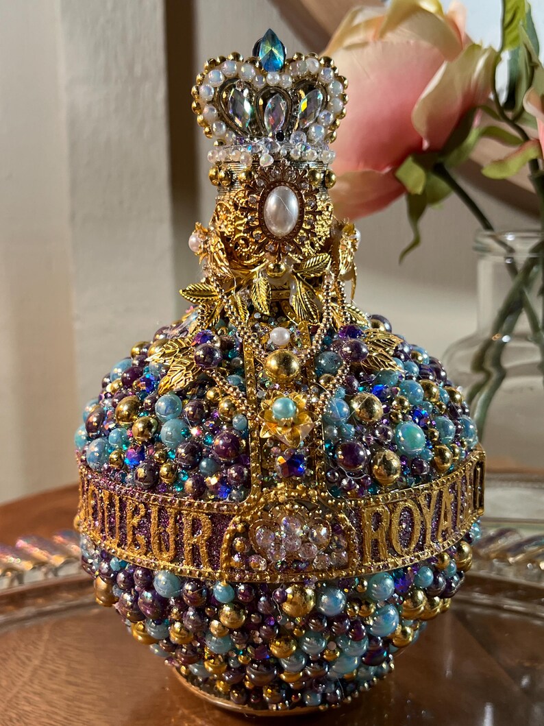 K&ouml;nnte beinhalten: Eine goldene und violette, mit Juwelen besetzte Flasche mit einem kronenf&ouml;rmigen Verschluss. Die Flasche ist mit Perlen, Strasssteinen und goldenen Akzenten verziert. Der Text "OUR ROYAL" ist um die Flasche herum geschrieben.