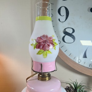 May include: A vintage oil lamp with a white frosted glass shade painted with pink flowers and green leaves. The lamp has a pink base and a brass top.