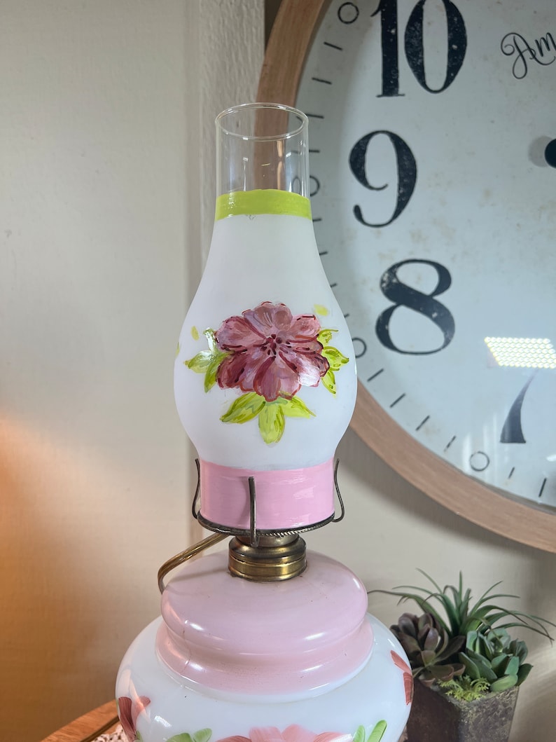 May include: A vintage oil lamp with a white frosted glass shade painted with pink flowers and green leaves. The lamp has a pink base and a brass top.