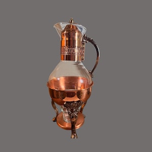 May include: A glass carafe with a copper neck and base, featuring a decorative metal handle and ornate feet. The copper neck has a textured band, and the glass body sits within a copper bowl supported by three detailed metal legs with claw feet.