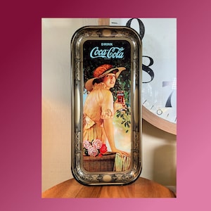 Puede incluir: Una bandeja de servir de metal vintage con un marco dorado ornamentado. La bandeja presenta un anuncio de Coca-Cola con una mujer con un vestido blanco sosteniendo un vaso de Coca-Cola.