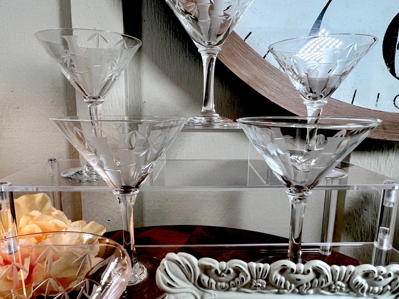 Op de afbeelding: Set van vier heldere glazen martini glazen met gegraveerde ontwerpen op een gelaagde acryl standaard. De glazen zijn gerangschikt in een piramidevorm.