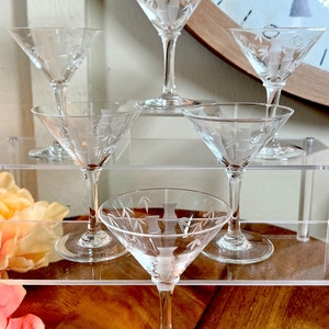 Op de afbeelding: Zes heldere glazen martini glazen met gegraveerde ontwerpen. De glazen zijn gerangschikt op twee heldere acrylplanken. De glazen zijn allemaal even groot en hebben dezelfde vorm, en ze hebben een delicaat, elegant ontwerp.