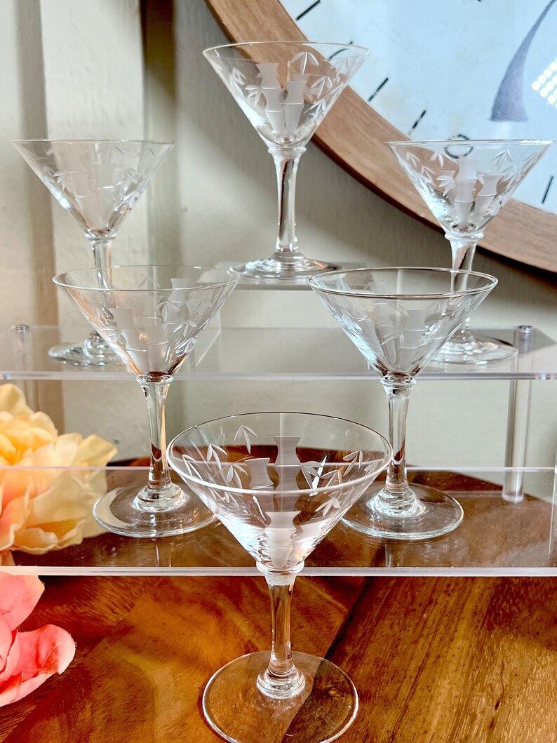 Op de afbeelding: Zes heldere glazen martini glazen met gegraveerde ontwerpen. De glazen zijn gerangschikt op twee heldere acrylplanken. De glazen zijn allemaal even groot en hebben dezelfde vorm, en ze hebben een delicaat, elegant ontwerp.