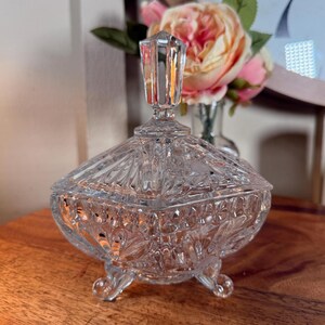 Pode incluir: Um recipiente para doces em vidro prensado transparente com corpo de padrão diamante, três pés pequenos e tampa a condizer com um remate pontiagudo facetado. A superfície de vidro reflete a luz, realçando as texturas geométricas complexas.