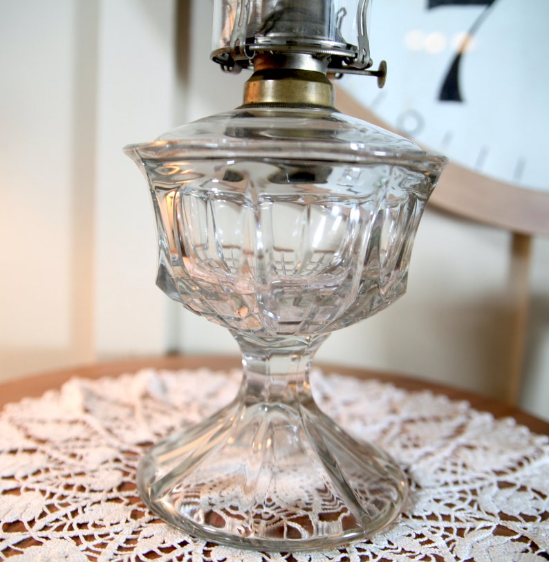 May include: A clear glass oil lamp with a cut glass design. The lamp has a metal top with a wick and a glass chimney.