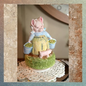 Puede incluir: Una figurita musical de cerámica de un cerdo con un vestido azul y amarillo y llevando dos cubos. El cerdo está de pie sobre una base verde con un pequeño lechón delante de él.