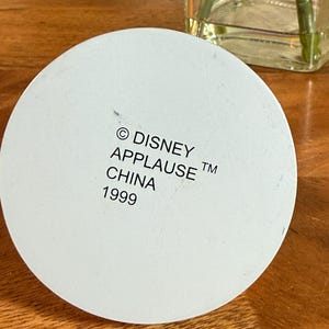 May include: A white, circular object with the text "&copy; DISNEY APPLAUSE TM CHINA 1999" printed on it. The object is set against a wooden surface, with a glass vase and plant visible in the background.