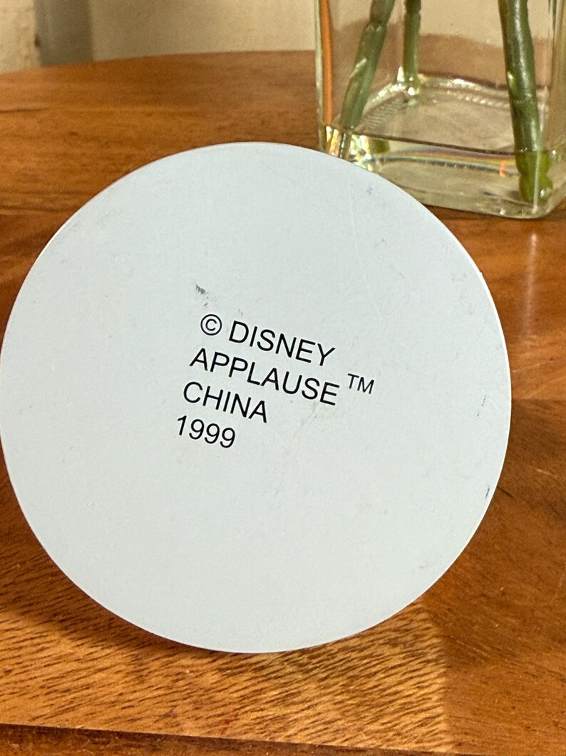 May include: A white, circular object with the text "&copy; DISNEY APPLAUSE TM CHINA 1999" printed on it. The object is set against a wooden surface, with a glass vase and plant visible in the background.
