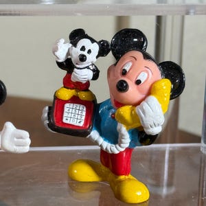 May include: A colorful Mickey Mouse figurine holding a yellow telephone receiver to his ear. The figurine is wearing a blue jacket, red pants, and yellow shoes. A smaller Mickey Mouse figurine is perched on a red telephone with white buttons. The figurine is a collectible item.