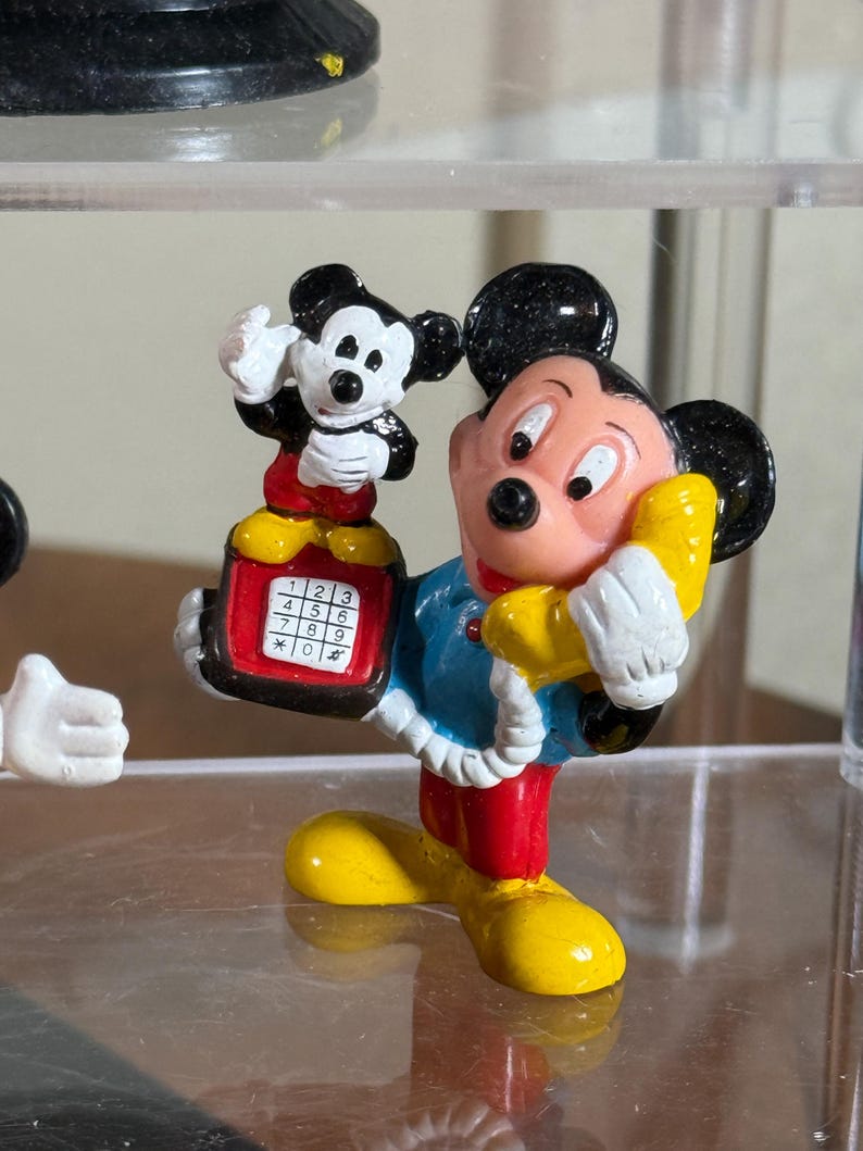 May include: A colorful Mickey Mouse figurine holding a yellow telephone receiver to his ear. The figurine is wearing a blue jacket, red pants, and yellow shoes. A smaller Mickey Mouse figurine is perched on a red telephone with white buttons. The figurine is a collectible item.
