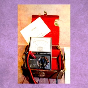 May include: A vintage black and silver analogue multimeter with a red and black probe wire, in a brown leather case with a red velvet lining. The case has a metal latch and a metal handle. The multimeter has a dial with the text "Bell & Howell Schools" and a needle that points to the "OFF" position. The multimeter is in a vintage leather case.