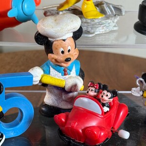 May include: A collection of Disney figurines, including Mickey Mouse as a chef, and Mickey and Minnie Mouse in a red car. Other figurines include a Mickey Mouse figure and a blue airplane toy. The figurines are on a clear surface.