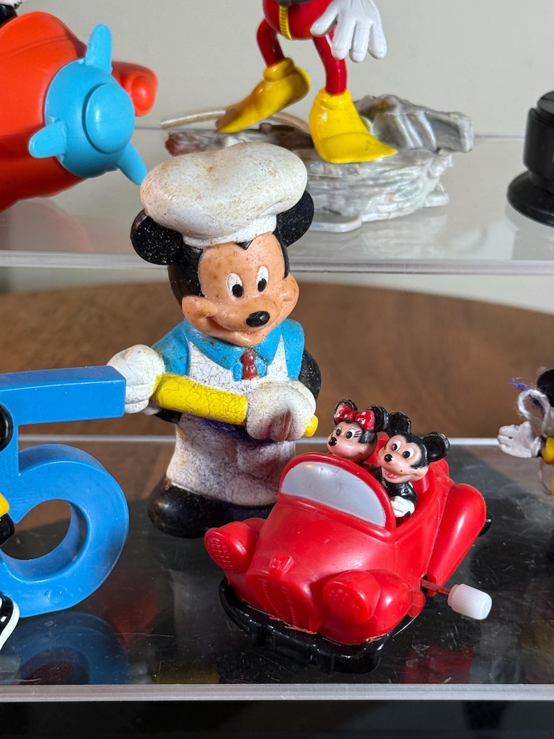 May include: A collection of Disney figurines, including Mickey Mouse as a chef, and Mickey and Minnie Mouse in a red car. Other figurines include a Mickey Mouse figure and a blue airplane toy. The figurines are on a clear surface.