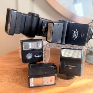 May include: Four black flash units for cameras. The flash units are labelled "Vivitar Autobounce 300 Dedicated", "Starblitz", "Quantarray MS-1", and "180".