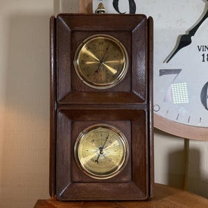 Puede incluir: Un conjunto de barómetro y termómetro de pared de madera con dos indicadores analógicos redondos. Los indicadores tienen caras doradas con marcas y manecillas negras. El indicador del barómetro dice "Barómetro" y el indicador del termómetro dice "Termómetro".