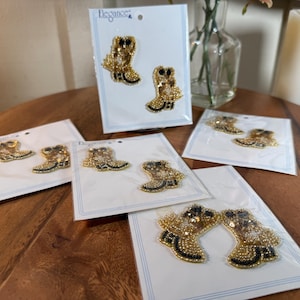 May include: Several pairs of gold and black beaded boot-shaped appliques on white cards. The appliques are embellished with sequins and beads. The cards are arranged on a wooden surface, with a few cards in the background.