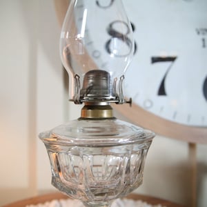 May include: A clear glass oil lamp with a decorative, faceted base and a metal chimney. The lamp has a brass burner and a clear glass globe.