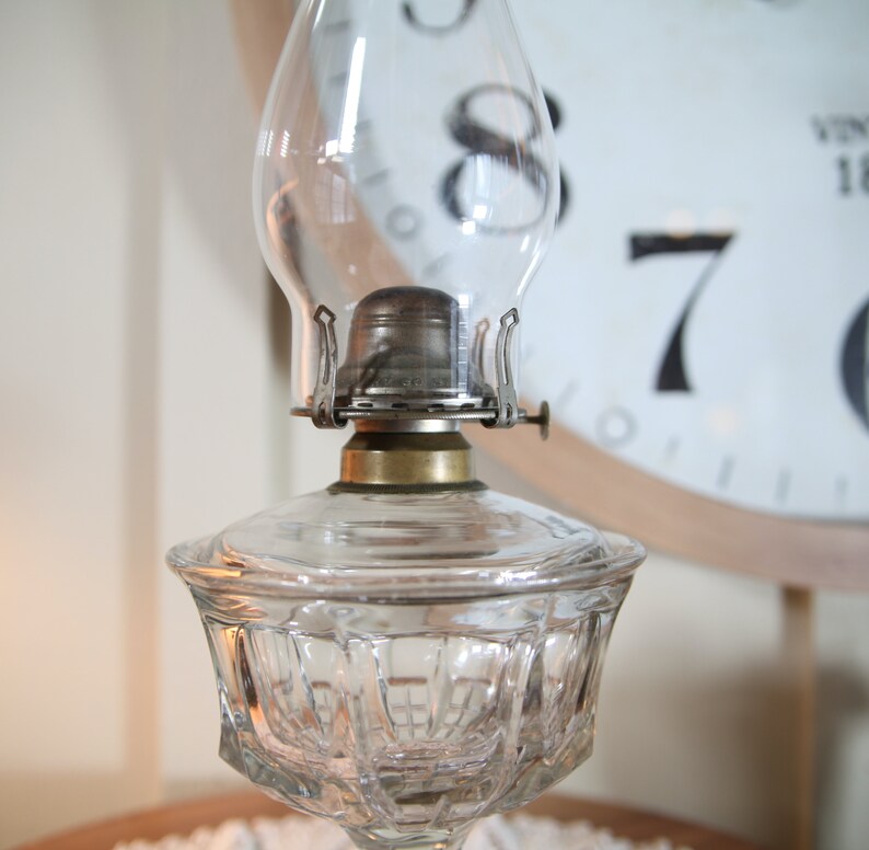 May include: A clear glass oil lamp with a decorative, faceted base and a metal chimney. The lamp has a brass burner and a clear glass globe.