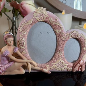 May include: A pink heart-shaped picture frame with a ballerina figurine. The frame has two oval openings and is decorated with ornate gold accents. The ballerina is in a seated pose, wearing a pink tutu.